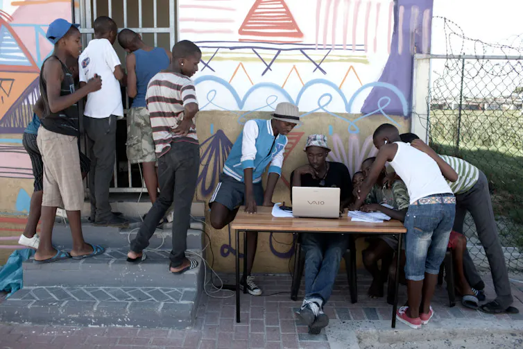 A group of boys gathers at a club house, one sitting and entering information into a computer at an outside table, others standing around him or peering through the security gate of the clubhouse, which sports painted murals of football players.
