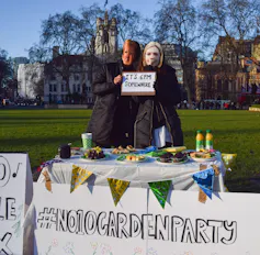 Two protesters wearing Boris and Carrie Johnson masks standing a table with party food on it holding a sign reading 'it's 6pm somewhere'