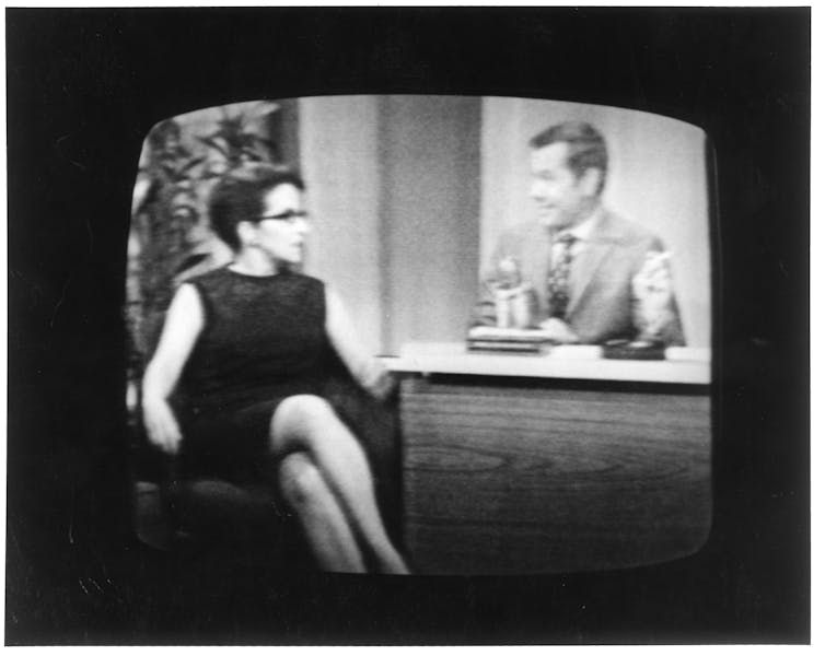 A black-and-white television screen shows a white woman sitting with her legs crossed as she is being interviewed by a man sitting behind a desk.
