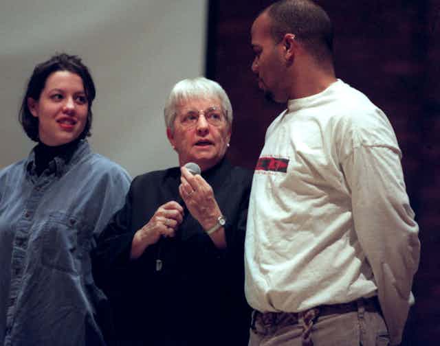 A gray-haired woman holding a microphone stands in between a Black man and a white woman.