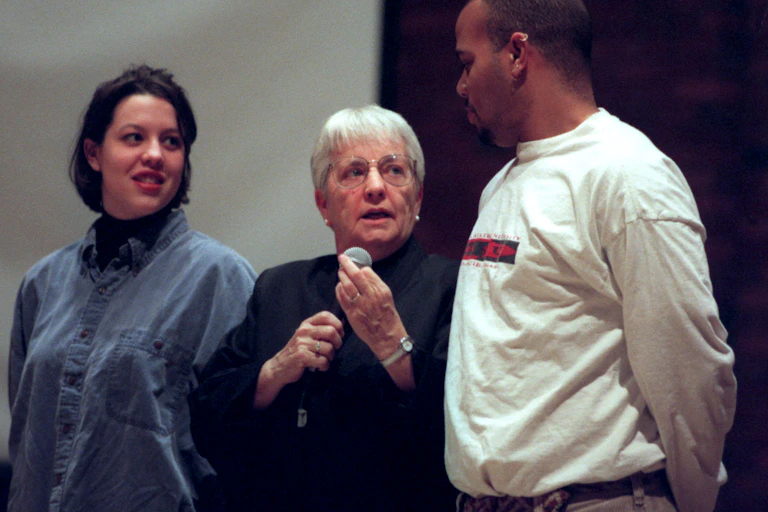 A gray-haired woman holding a microphone stands in between a Black man and a white woman.