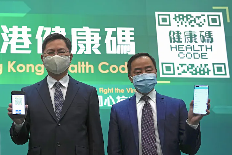 two Chinese officials hold up mobile phones as they stand in front of a green background with a QR code
