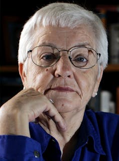 A woman with gray hair and wire-rimmed glasses rests her chin on her hand.