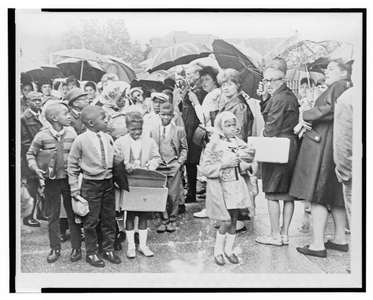 A black-and-white photograph shows Black schoolchildren with book bags and lunchboxes walking past a line of white adults, many holding umbrellas.