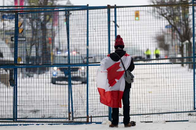 A person stands draped in a flag facing a fenced-off area.