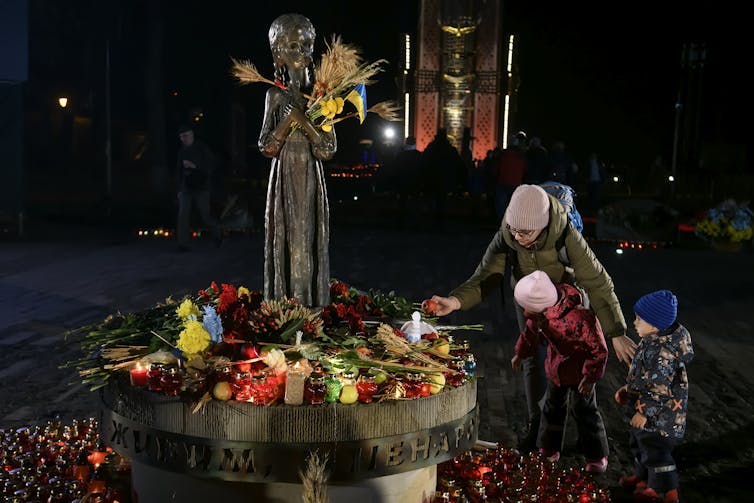 A woman and children, dressed for the cold weather and during the night, leave a piece of fruit at a monument.