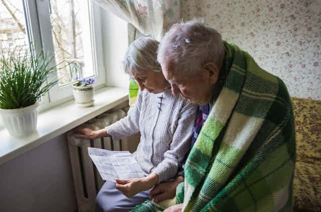 An elderly couple huddled around a heater in a blanket