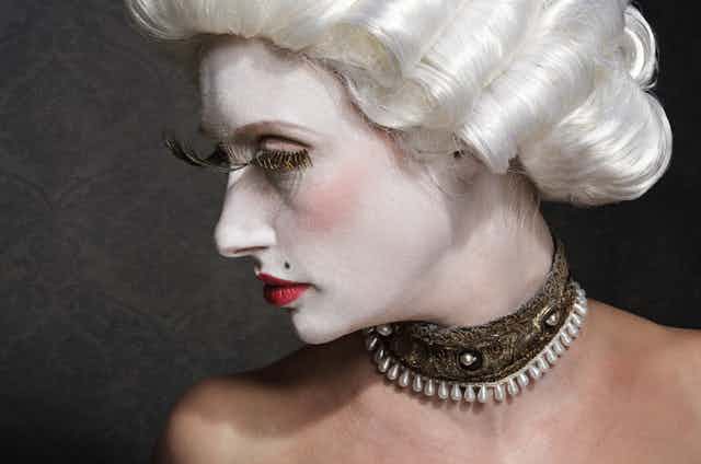 An actor wearing white make-up, a white wig, exaggerated false eyelashes and a costume pearl and lace choker