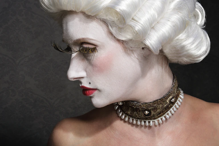 An actor wearing white make-up, a white wig, exaggerated false eyelashes and a costume pearl and lace choker