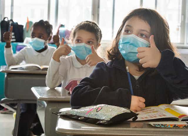 Three girls sitting at school desks, wearing blue face masks and giving a thumbs-up sign