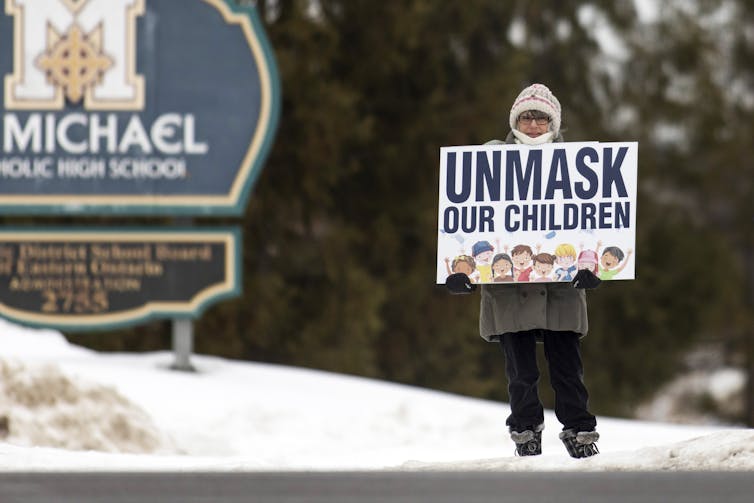 A protesting in winter clothes holds a sign reading'Unmask our children' outside a school
