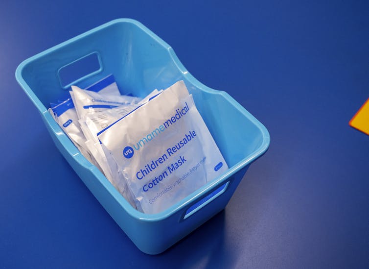 A blue plastic container filled with individually wrapped children's size cotton face masks, on a blue table