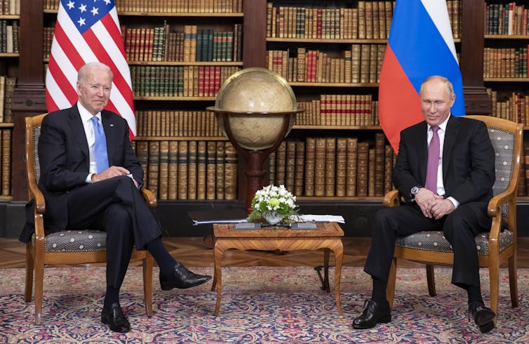 Joe Biden and Vladimir Putin seated in armchairs in front of their respective country flags, in a library with a globe and small table between them