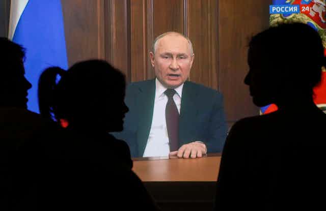 Vladimir Putin makes a televised speech from the Kremlin watched by people shown here in silhouette.