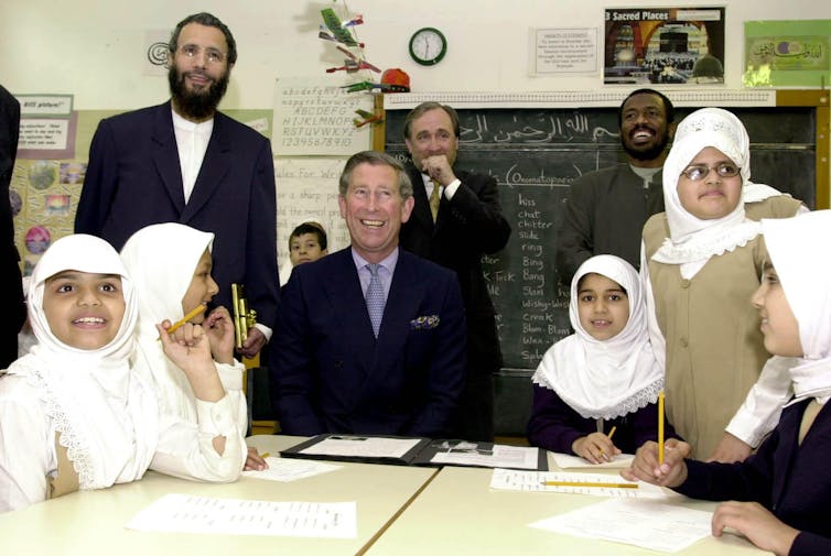 The Prince of Wales is seated at a school table with smiling little girls in white headscarves and teaching staff