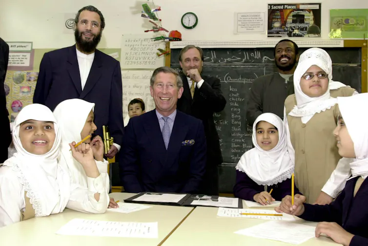 The Prince of Wales is seated at a school table with smiling little girls in white headscarves and teaching staff