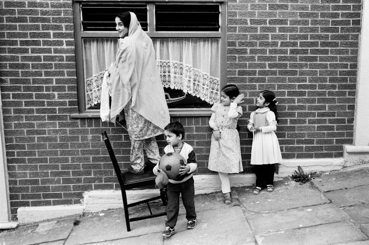 A mother in a salwar kameez stands on a chair on the pavement with children playing around her.