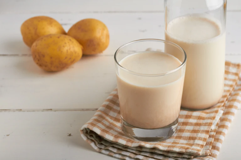 A glass and jug of potato milk on a tea towel, with three potatoes on the table behind it.