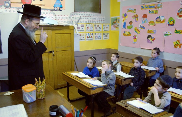 A rabbi in a black coat and hat speaks to a classful of small boys in yarmulkes.