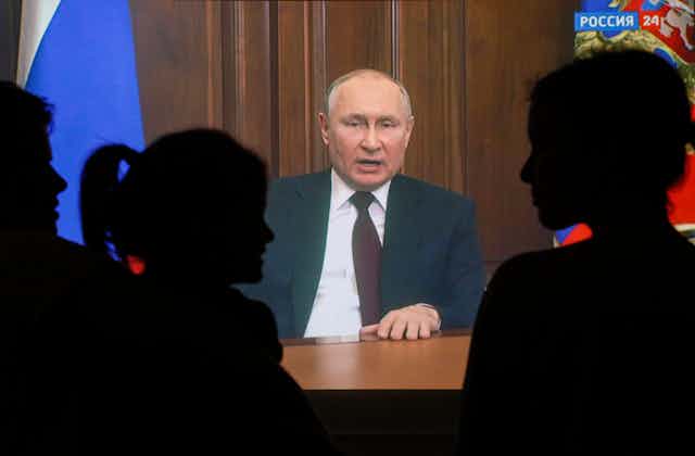 Vladimir Putin pronuncia un discurso televisado desde el Kremlin, observado por personas que aparecen aquí en silueta.