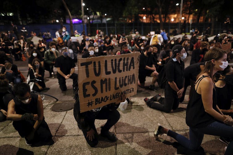 A kneeling man in a crowd holds a sign reading #BlackLivesMatter and 'tu lucha es mi lucha' (your fight is my fight).