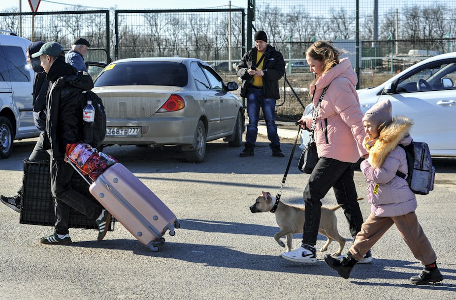 Side view of people walking in a parking lot carrying bags and suitcases, one woman walks a dog and holds her child's hand