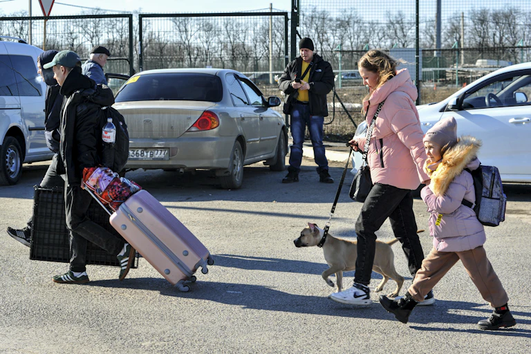 Side view of people walking in a parking lot carrying bags and suitcases, one woman walks a dog and holds her child's hand