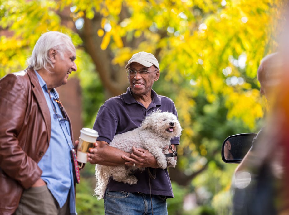 Two elderly man in a conversation while walking in a park, one holding a pet dog.