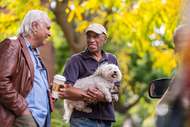 Two elderly man in a conversation while walking in a park, one holding a pet dog.