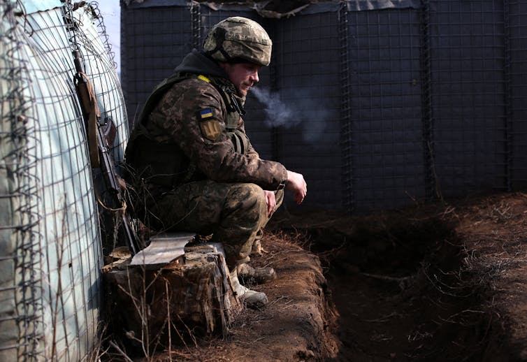 A soldier wearing military garb, with a Ukrainian flag on the uniform, sits and smokes in a mud trench.