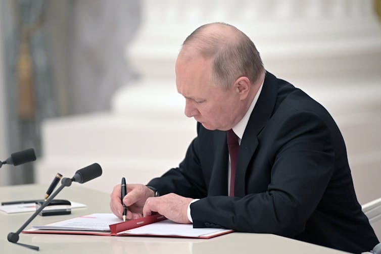 A man dressed in a suit sits behind a white desk and uses a pen to sign papers.