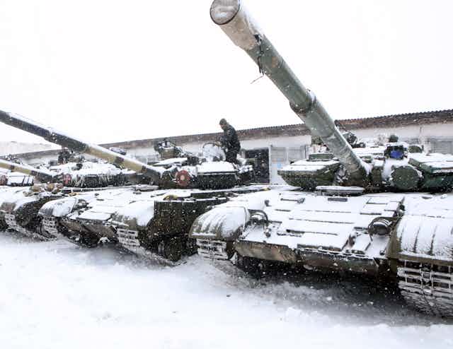Battle tanks with their cannons pointed toward the sky are parked in a row on a snow-covered patch of land.