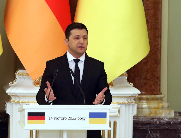 A man in a dark suit stands behind a lectern and is answering questions from reporters.