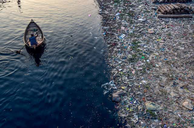 A man paddles a boat along a river whose bank is lined with plastic trash