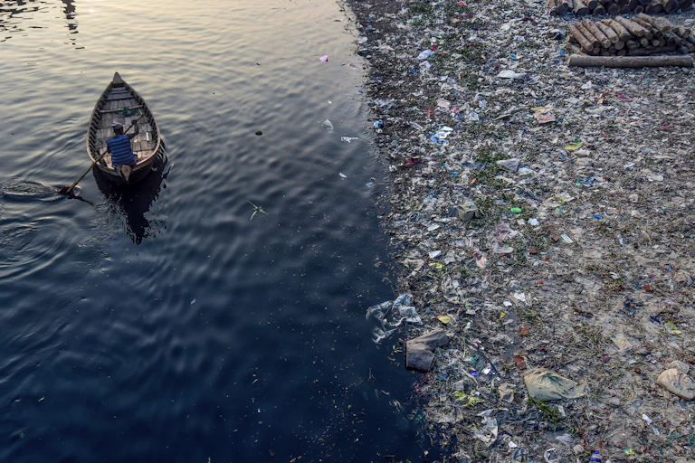 A man paddles a boat along a river whose bank is lined with plastic trash