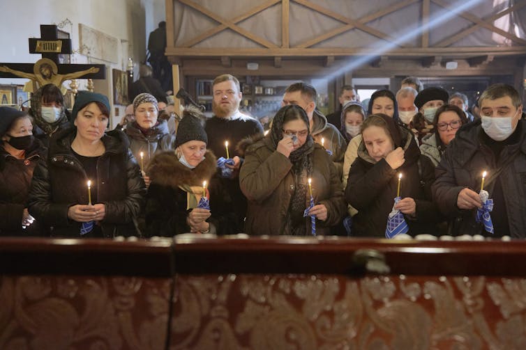 Men and women dressed in black and brown are gathered in two lines holding candles.