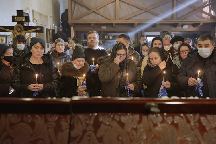 Homens e mulheres vestidos de preto e marrom estão reunidos em duas filas segurando velas.