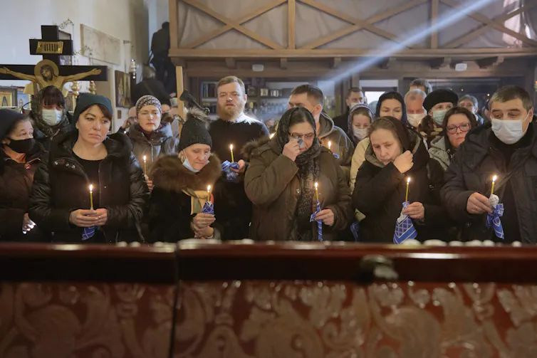 Men and women dressed in black and brown are gathered in two lines holding candles.