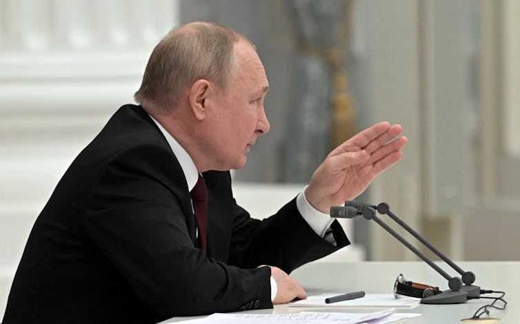 Russian President Vladimir Putin, wearing black jacket and white shirt, sitting at a table and talking to a meeting in a large hall.