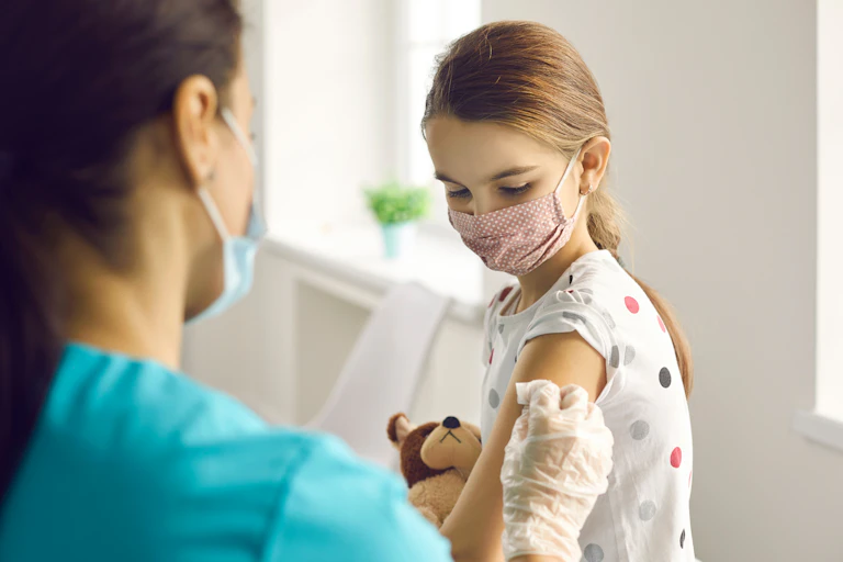 Nurse immunises child in a mask, holding a teddy.