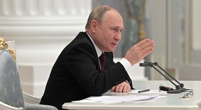 profile view of Vladimir Putin sitting at a desk and speaking into a microphone while gesturing with his right hand