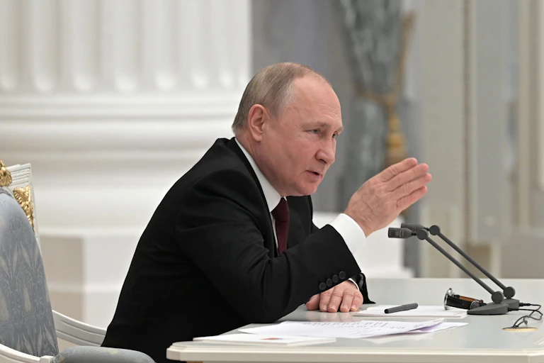 profile view of Vladimir Putin sitting at a desk and speaking into a microphone while gesturing with his right hand