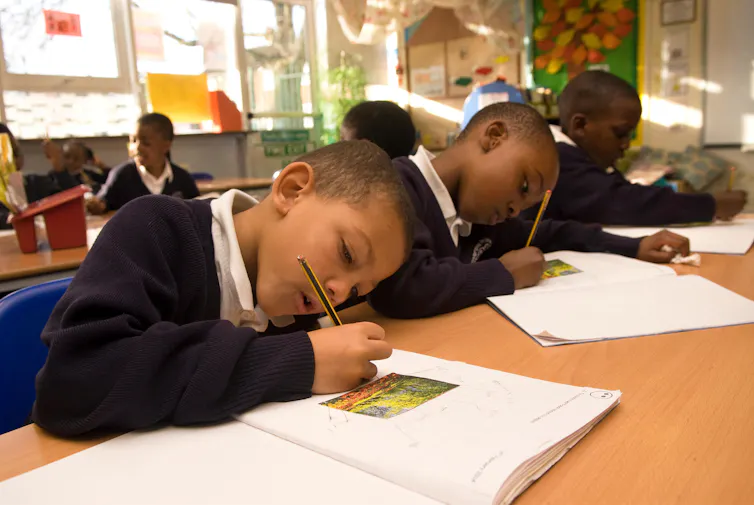 Three black boys in school uniform work at a desk with pencils.