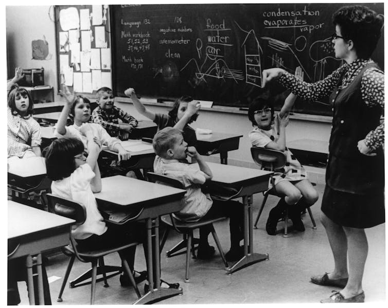 A white woman stands by a classroom blackboard in front of white students sitting at desks, many with their hands raised.