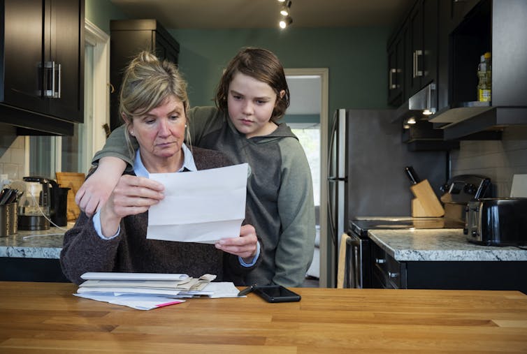 A woman seated in a kitchen looks over paperwork while a boy lingers over her shoulder with his arm around her.