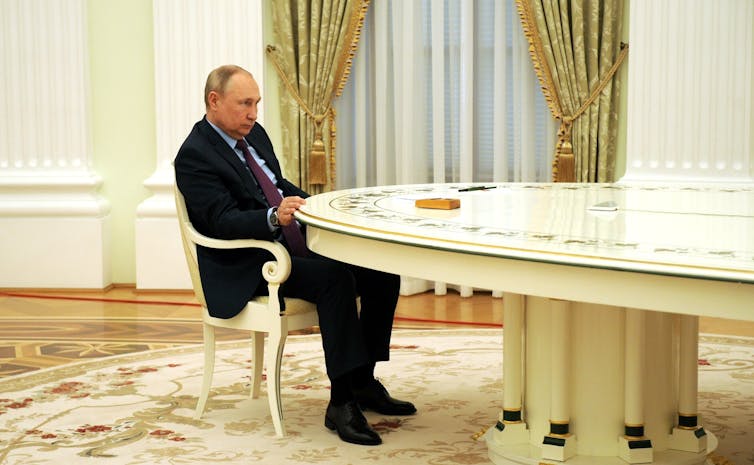 Russian President Vladimir Putin in a dark suit looking serious as he sits at the head of a very big table.