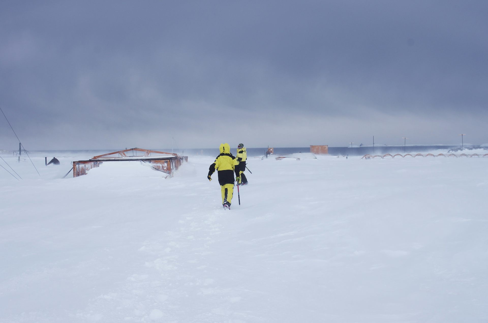 Two researchers walking on ice at Wilkes Station next to an abandoned building buried in the ice.
