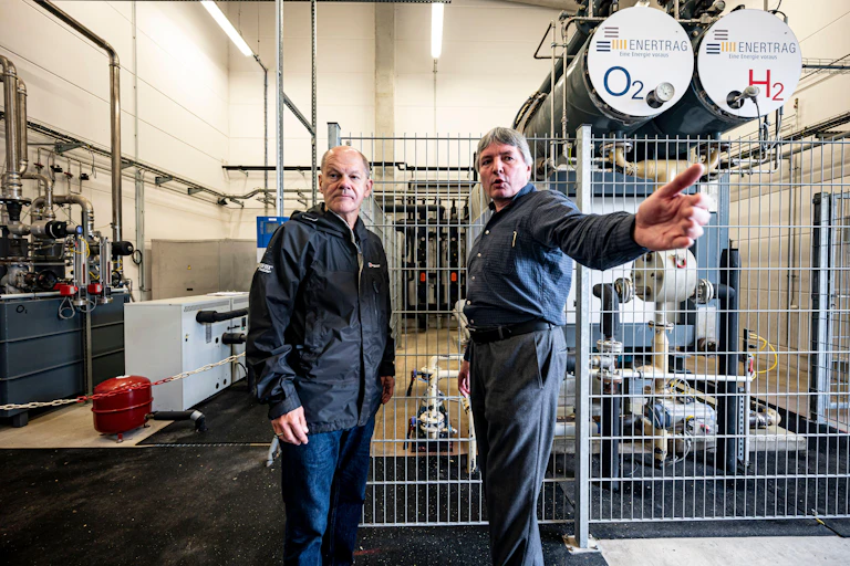 Two men stand inside a power plant with surrounded by machinery.
