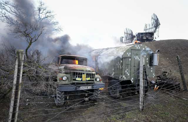 Damaged military equipment, some on fire and sending smoke into the air, behind a post and wire fence.
