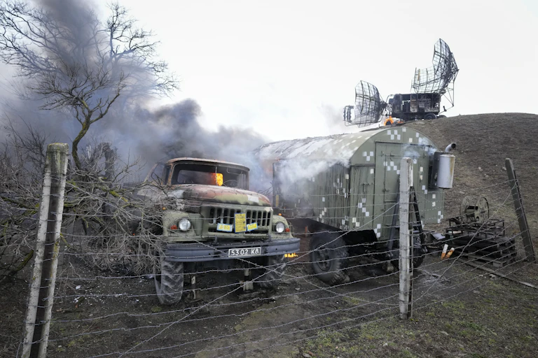 Damaged military equipment, some on fire and sending smoke into the air, behind a post and wire fence.